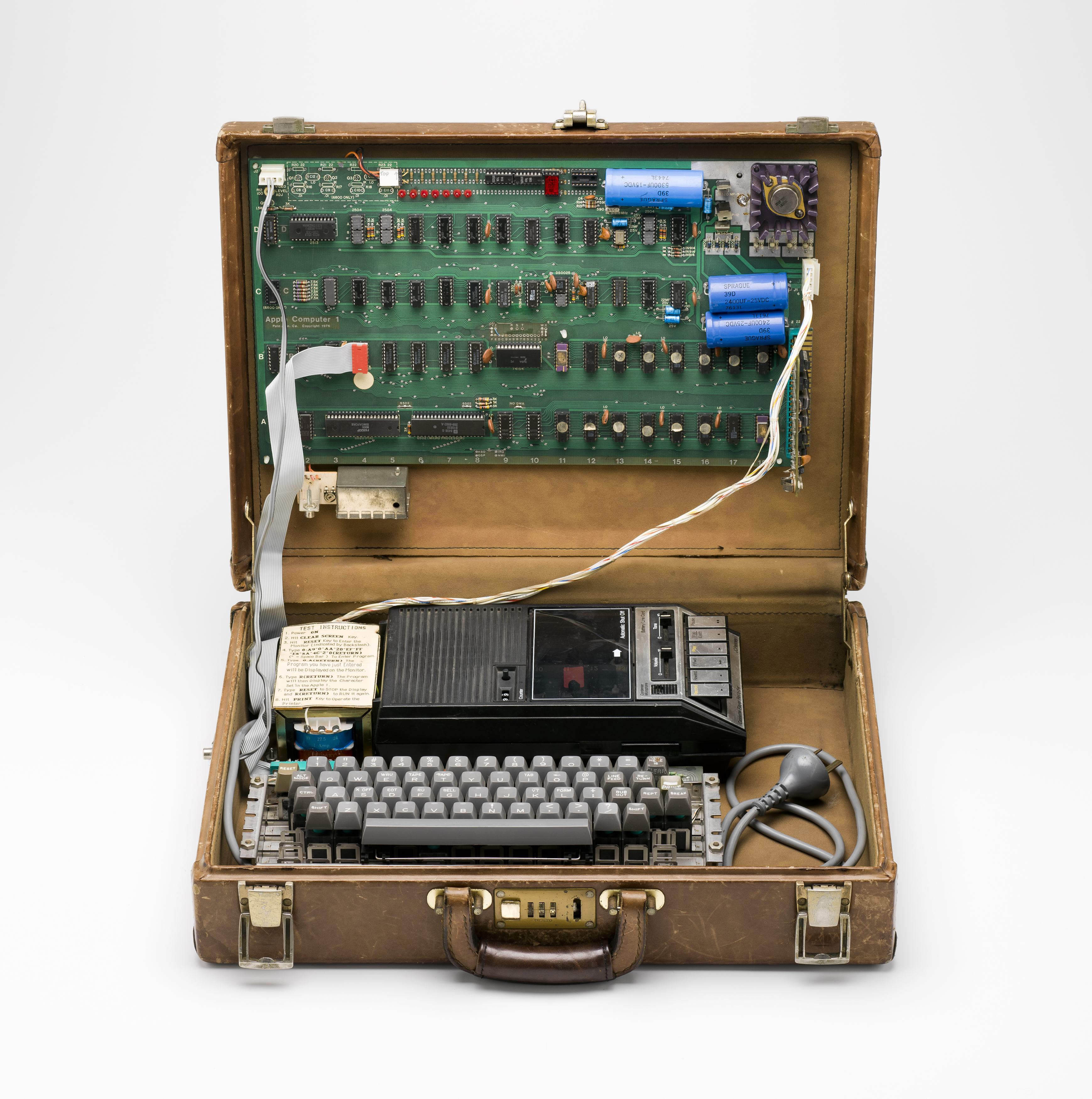 A brown leather suitcase is open withe the green motherboard attached to the inside of the lid. Sitting in the suitcase base is a keyboard and tape recorder.